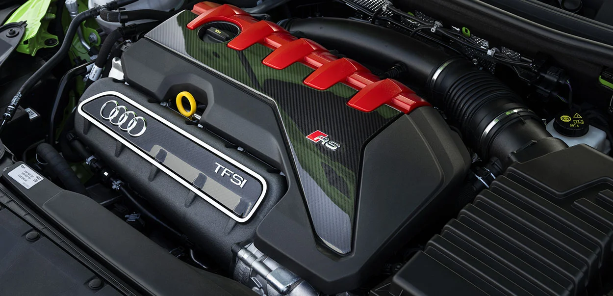 Moteur Audi Sport 5-cylindres 2.5 TFSI sous le capot, montrant le cache moteur en fibre de carbone et la rangée de bobines rouges, avec logos Audi et RS visible
