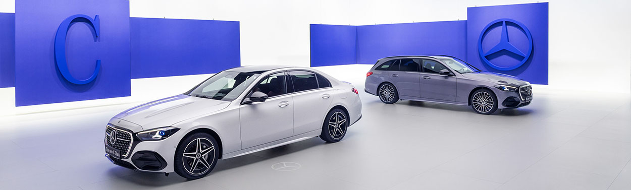 La nouvelle Mercedes-Benz Classe C Berline argentée et le Break gris 2026 sur un stand d'exposition avec des logos de la marque en bleu.