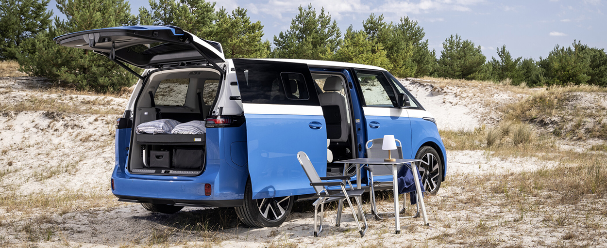 Volkswagen ID. California Cruise bleu et blanc avec coffre ouvert sur un espace couchage et mobilier de camping installé