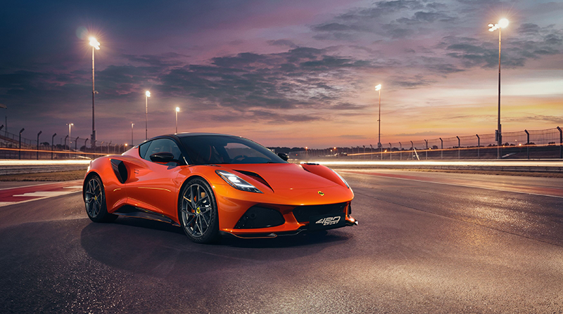 alt="Vue de trois-quarts face d'une Lotus Emira 420 Sport orange sur un circuit automobile éclairé au coucher du soleil