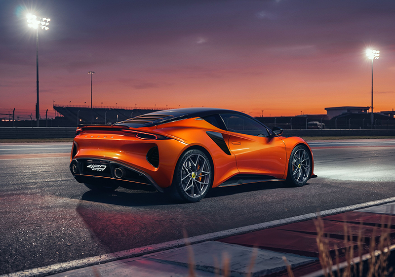 alt="Vue arrière de trois quarts d'une Lotus Emira 420 Sport orange métallisé sur une piste de course au crépuscule, avec diffuseur et échappements visibles."