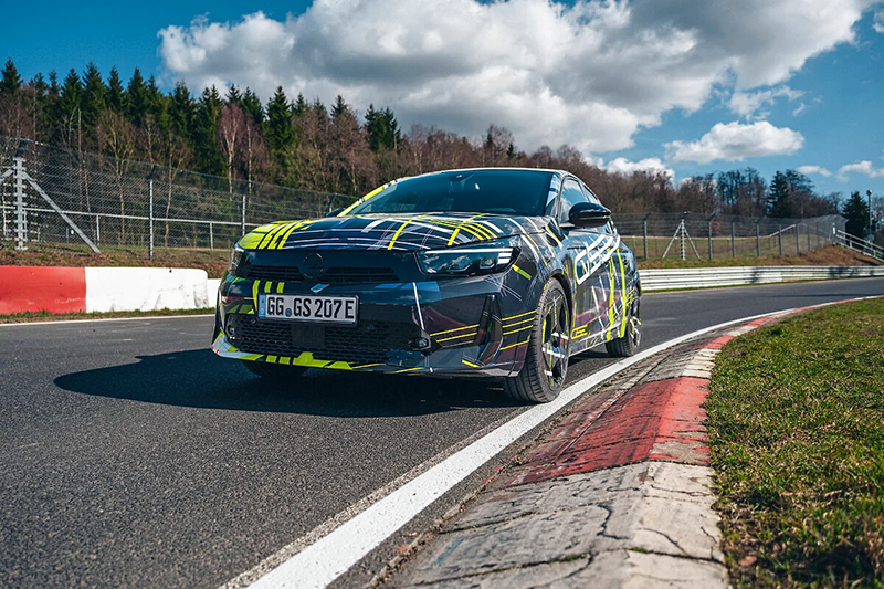 Nouvelle Opel Corsa GSE 2027 en test de performance sur circuit, livrée camouflage noir et jaune avec motifs géométriques rétro.