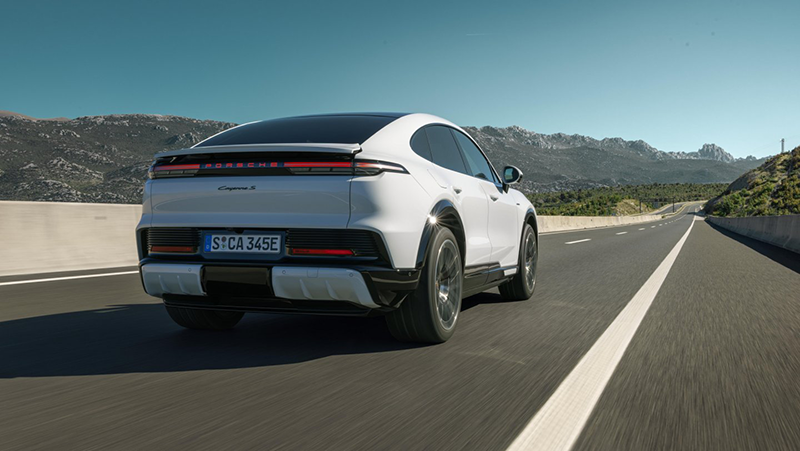 Vue arrière dynamique du Porsche Cayenne Coupé Electric en mouvement sur route, mettant en avant son aileron adaptatif déployé et son bandeau lumineux LED.