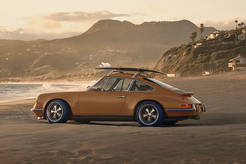 Porsche 911 Singer orange avec planches de surf sur une plage de Californie au soleil couchant.